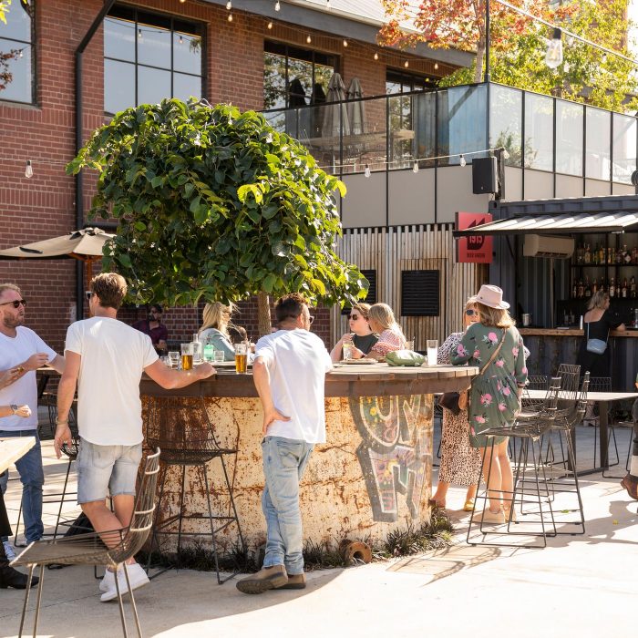 A lively outdoor 1915 beer garden scene with people gathered around a circular bar that has a large tree in the center. The bar is in 1915's industrial-style setting with brick buildings and metal accents. Patrons are casually dressed, enjoying drinks and conversations. A man is seen gesturing with two young children nearby, while others are seated or standing around the bar. The atmosphere is relaxed and sunny, with greenery and string lights adding to the ambiance.