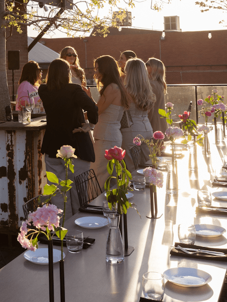 Rooftop event at sunset with a group of people gathered near a rustic bar, surrounded by a long dining table decorated with colorful floral arrangements in slender vases. Plates, glasses, and utensils are neatly set, creating a warm and stylish ambiance against the backdrop of a sunlit brick building and string lights.