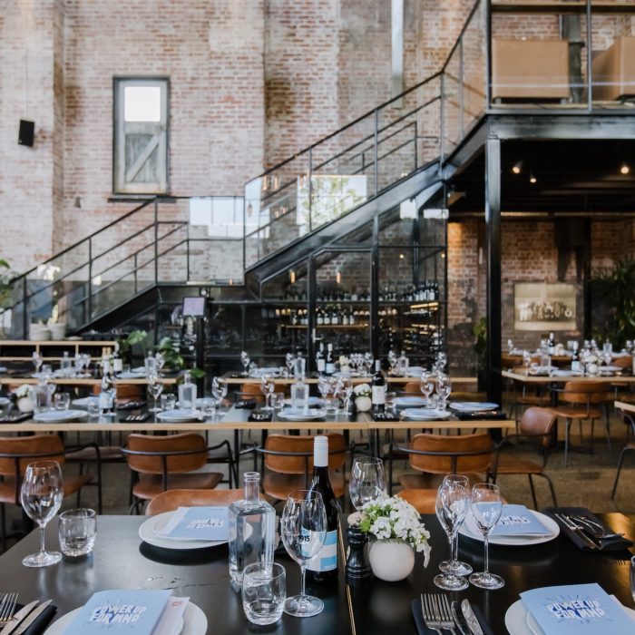 Elegant dining setup in an industrial-style event space with exposed brick walls, high ceilings, and large glass-enclosed staircase. Tables are set with plates, wine glasses, bottled water, and small floral arrangements, creating a refined, welcoming atmosphere.